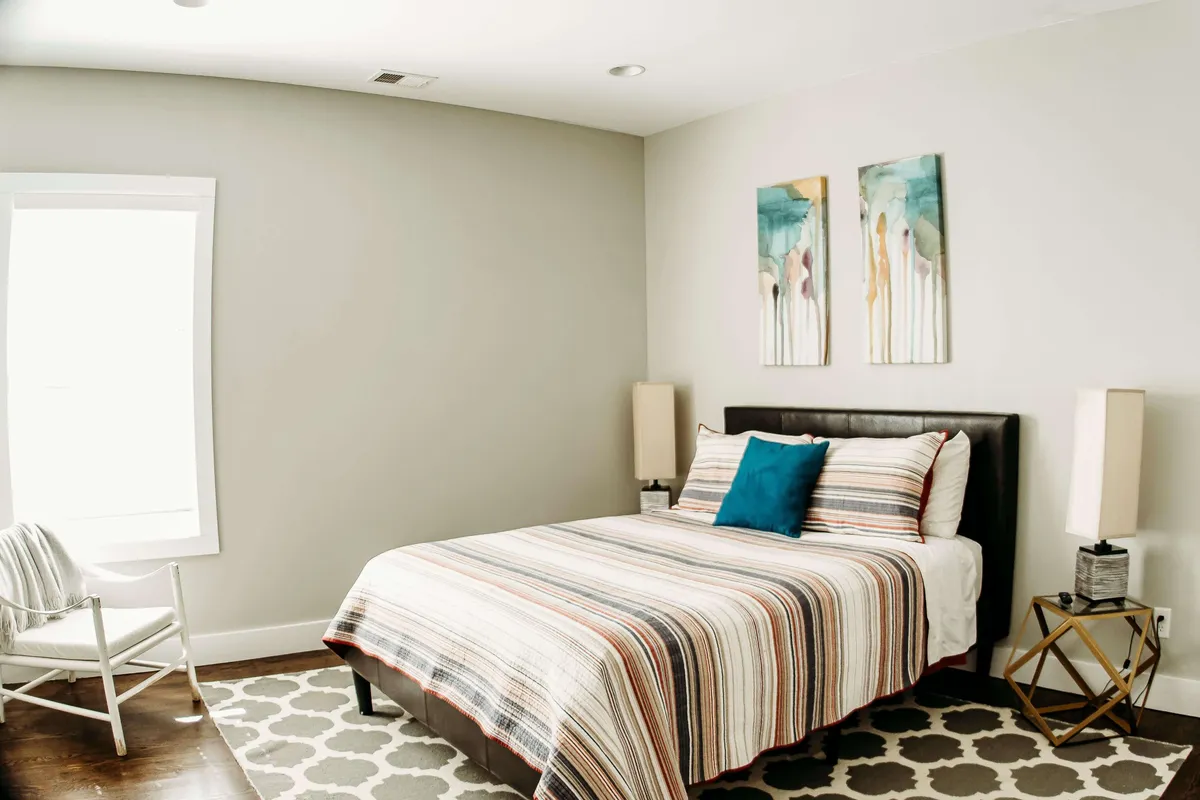 A serene bedroom featuring a bed with striped bedding, accent pillow, decorative side table, and calming artwork on the wall. The mood is tranquil and cozy, making it an inviting space to unwind after a busy day.