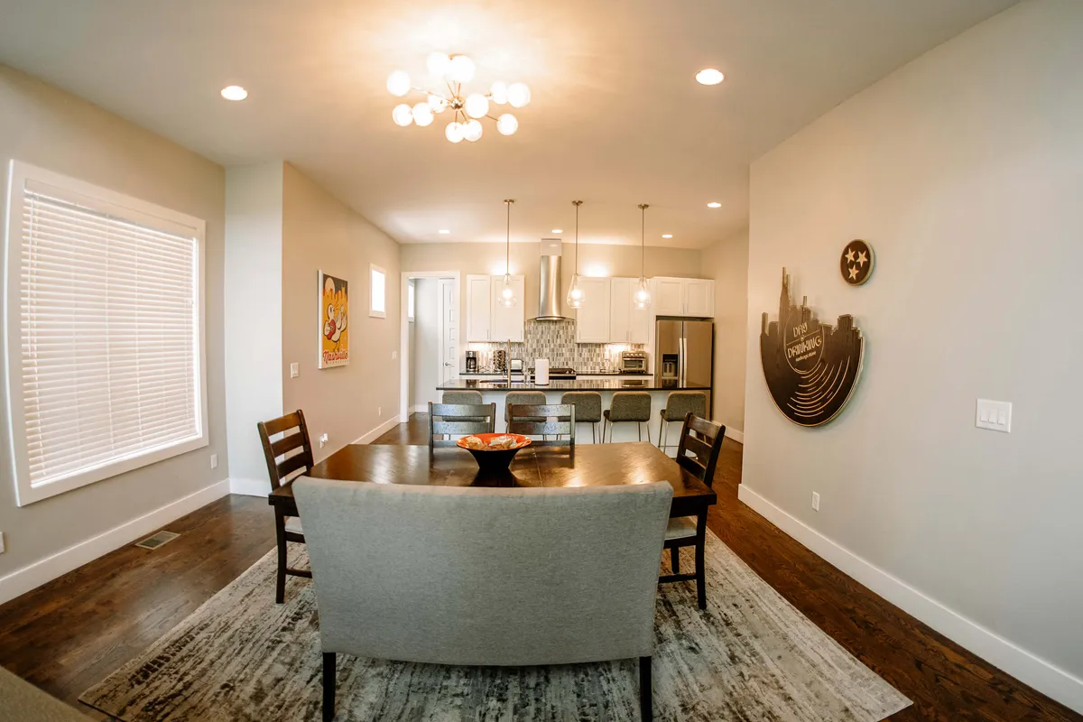 The photo highlights a large dining table beneath a chandelier, with the kitchen in the background and wall art providing a cozy, elegant atmosphere. The setup is ideal for family-style meals, holiday celebrations, or intimate dinner parties with friends.