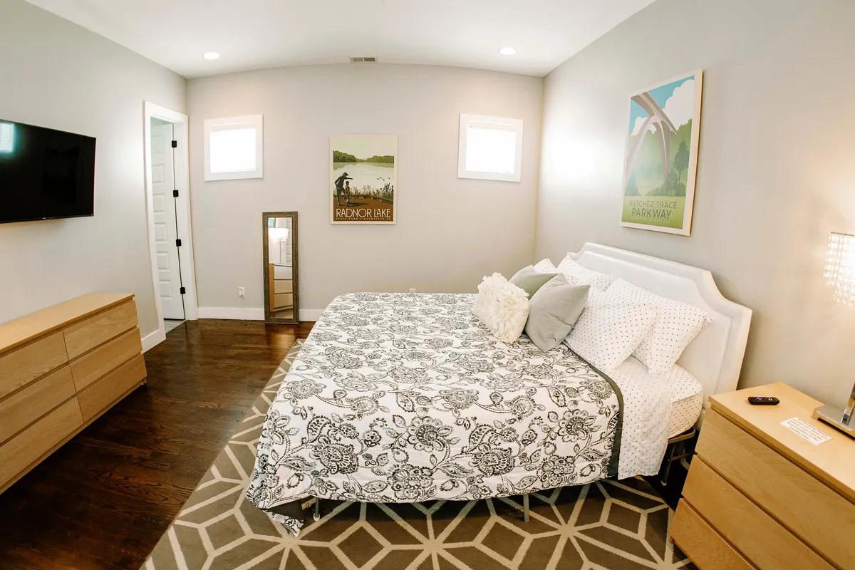 A cozy bedroom with patterned bedding, a wall-mounted TV, a mirror, a nightstand with decorative items, and calming wall art. Soft natural light fills the room, highlighting the serene environment.