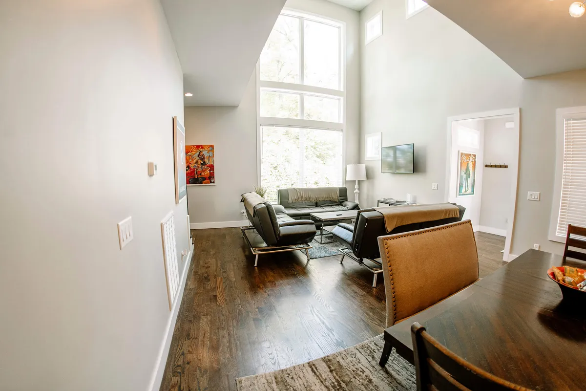 A contemporary living room with large windows, modern furniture, and artwork on the wall creates a cozy yet stylish setting for relaxation or entertaining. The room is filled with natural light and has a welcoming vibe for lounging, watching TV, or casual conversation.